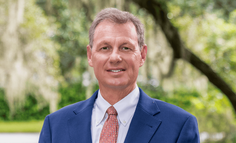 Smiling attorney in a navy suit, white shirt, and red tie, photographed outdoors against blurred live oaks and Spanish moss.