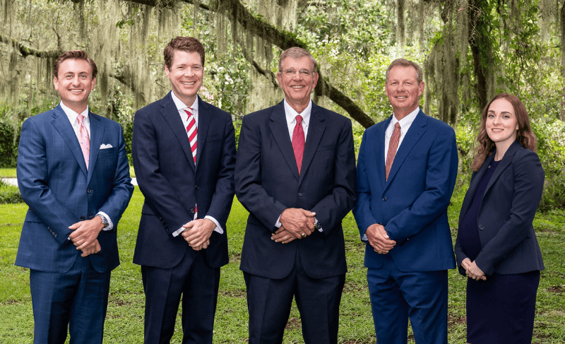 Five attorneys, four men and one woman, in navy and dark suits stand side by side on a lawn beneath live oaks draped in Spanish moss, smiling with hands clasped.