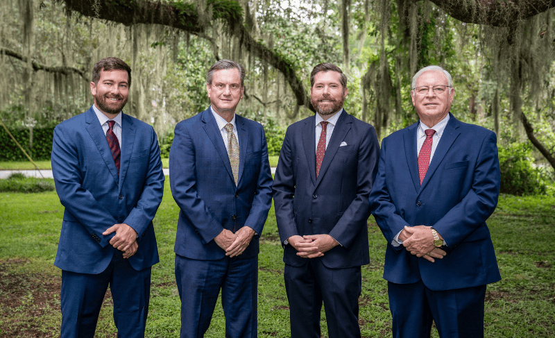 Four attorneys in navy suits stand side by side on a green lawn beneath live oaks draped in Spanish moss, smiling with hands clasped in front of them.